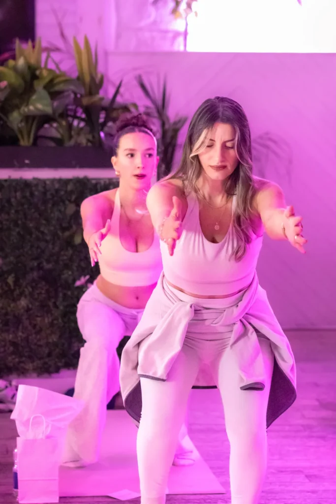 Two women in neutral activewear squatting with arms reaching forward in a group Pilates class